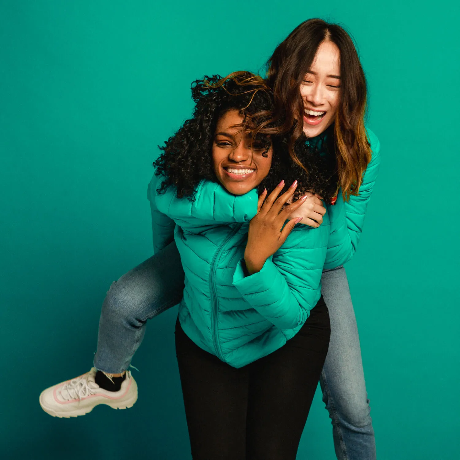 Black woman piggy backing Asian woman on a green background